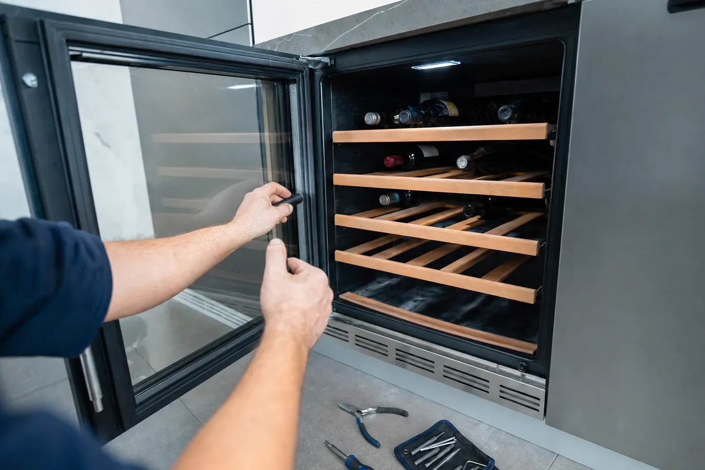 Hands at the open glass door of an undercounter wine cooler, pliers and a driver roll on the floor alongside