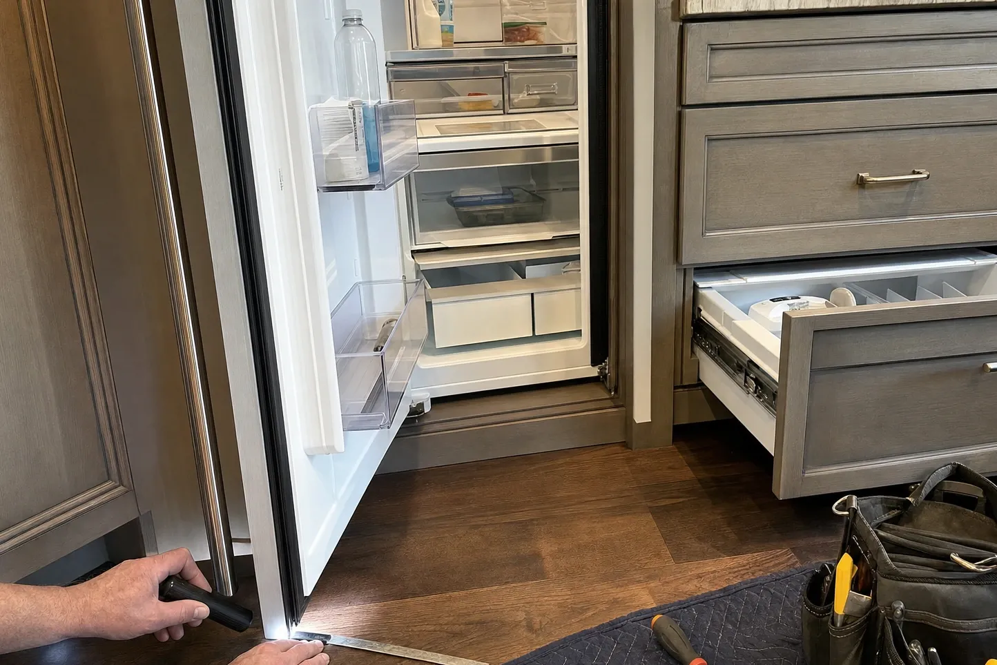 Panel-ready built-in refrigerator standing open in a fitted kitchen, a hand holding a flashlight and steel rule at the door edge