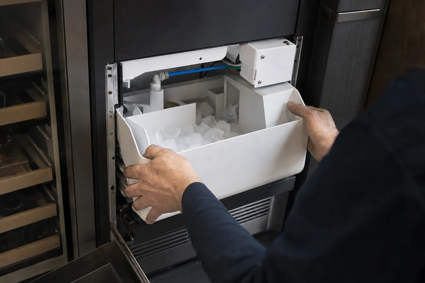 Hands lifting a white ice bin of clear cubes out of a built-in ice maker, a wine cooler with wooden racks alongside