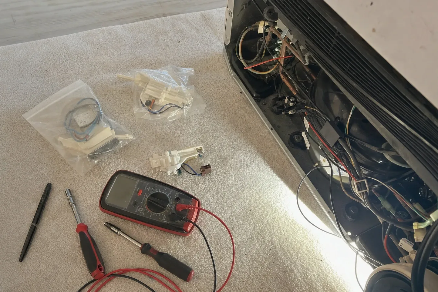 Multimeter, nut drivers and bagged replacement parts laid out on the floor beside an opened machine compartment
