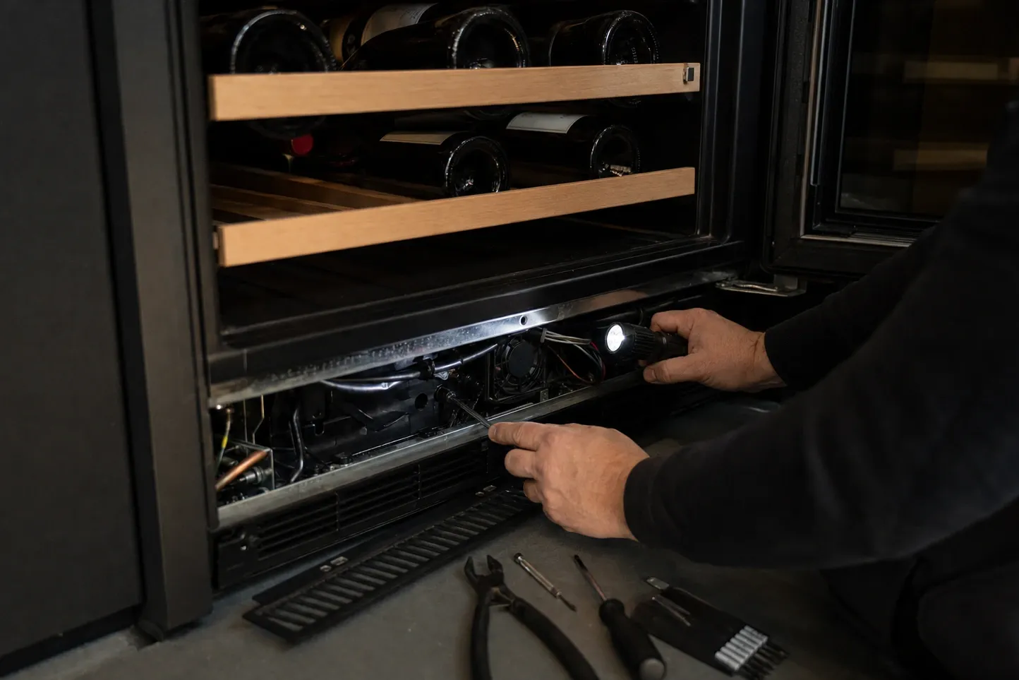 Base grille lifted off an undercounter wine unit and laid on the floor, a torch lighting the small fan and wiring inside the plinth